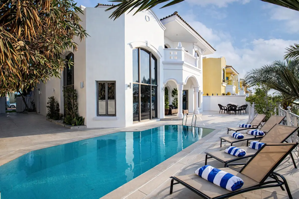 A wide angle view of the villa's backyard featuring a lush green lawn, a stone patio with outdoor seating, and a direct view of the open water.